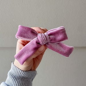 Puede incluir: Un lazo para el pelo de terciopelo malva con un acabado ligeramente brillante en toda la tela. El lazo está construido con un detalle de nudo central y dos bucles horizontales que se extienden hacia los lados, mostrando una apariencia suave y texturizada.