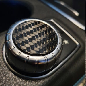 May include: Close-up of a silver knob with a black carbon fiber insert. The knob has a textured surface and is surrounded by a black leather surface.
