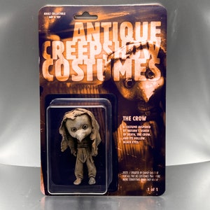 May include: A collectible figure of a crow-like creature with a beak and hollow eyes. The figure is wearing a tattered brown robe and is posed standing. The packaging features the text "Antique Creeps Show Costumes" and "The Crow".