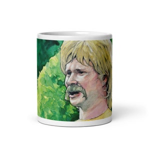Puede incluir: Una taza de cerámica blanca con una ilustración de acuarela de una persona con cabello rubio y bigote. La persona lleva una camisa amarilla y está rodeada de follaje verde.