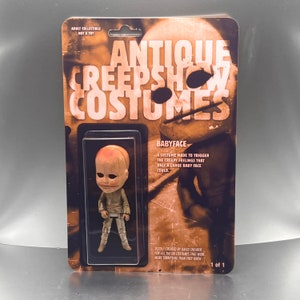 May include: A plastic package with a brown and white background featuring a small, pale, doll-like figure with a large head and a creepy expression. The package is labeled "Antique Creeps Show Costumes" and "Babyface". The text reads "A costume made to trigger the creepy feelings that only a large baby face could."