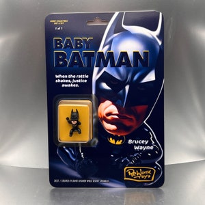May include: A black and yellow card featuring a cartoon image of a baby Batman with the text "Baby Batman" and "When the rattle shakes, justice awakes." The card also features the text "Brucey Wayne" and "ReWork Toys".