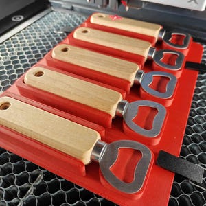 Op de afbeelding: Een rode plastic houder met zes houten flessenopeners. Elke opener heeft een metalen flessenopener aan het uiteinde. De houder staat op een grijs honingraat oppervlak.