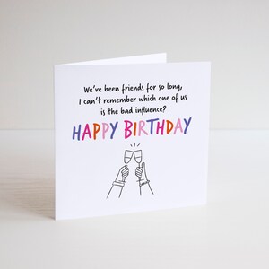 May include: A white card with a humorous birthday message that reads "We've been friends for so long, I can't remember which one of us is the bad influence? Happy Birthday". The card features a simple line drawing of two hands raising glasses of champagne.
