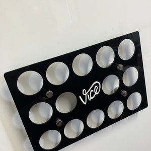 May include: A black acrylic organizer with 15 circular openings. The word "Vice" is written in white script. The organizer is mounted with silver screws against a white background. The design is angled.