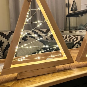 Puede incluir: Decoración de árbol de Navidad de madera con luces de hadas blancas cálidas dentro del marco triangular. El árbol es de madera clara y se apoya sobre una base de madera. Las luces están espaciadas uniformemente.