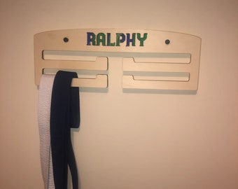 Könnte beinhalten: Ein Gürtelhalter aus Holz mit dem Namen "RALPHY" in blauen und grünen Buchstaben. Der Halter hat Schlitze zum Halten von Gürteln, und zwei Gürtel hängen an dem Halter. Der Halter ist an einer beigefarbenen Wand befestigt.