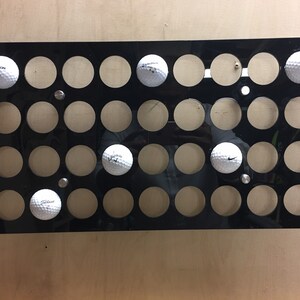 May include: A black acrylic golf ball display case with 30 circular openings. Several white golf balls with brand logos are visible in the openings. The case is mounted on a light wood surface.