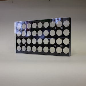 May include: A rectangular black acrylic display with multiple rows of evenly spaced, circular cutouts. The clear acrylic front allows visibility of the circles. The display is mounted with silver hardware and is set against a white background.