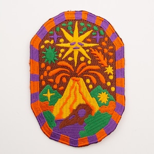 Puede incluir: Parche bordado con un diseño colorido que presenta un volcán, un sol y varios elementos celestiales y naturales. El parche tiene un fondo marrón con detalles en naranja, amarillo, verde y morado. El borde es naranja y morado.