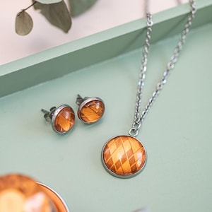 May include: A jewelry set featuring a necklace and matching stud earrings. The necklace has a silver chain and a round pendant with a brown and orange diamond pattern. The earrings and pendant have a silver bezel.