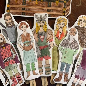 Puede incluir: Un conjunto de 10 muñecas de papel pintadas a mano con mangos de palo. Las muñecas están vestidas con varios disfraces, incluyendo un vikingo, una reina y un señor. Las muñecas están dispuestas frente a un cofre de madera.