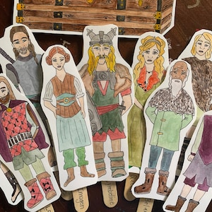 Puede incluir: Un conjunto de muñecas de papel que representan personajes de una obra de teatro o una historia. Las muñecas están pintadas a mano y presentan trajes y expresiones detalladas. Las muñecas están dispuestas sobre un fondo de madera con un cofre de madera en el fondo. Las muñecas están etiquetadas con nombres como "Imogen", "Ymbeline" y "Belarius".