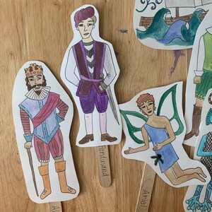 May include: Three paper puppets with stick handles. The puppets are a king with a crown, a man in a purple and black costume, and a fairy with green wings. The king puppet has the text "Ferdinand" on the stick handle. The fairy puppet has the text "Ariel" on the stick handle.