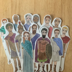 May include: A set of 12 paper cutouts of people in various poses and clothing. The figures are drawn in a cartoon style and are all wearing different outfits, including a toga, a tunic, and a dress. The figures are all standing on a wooden surface.