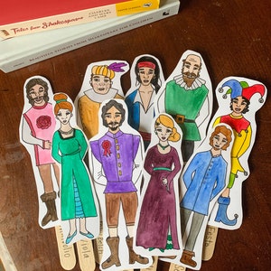 May include: A set of ten paper puppets depicting characters from Shakespeare's plays. The puppets are hand-drawn and colored with watercolors. Each puppet has a stick attached to the back for holding. The puppets are arranged in a semi-circle on a wooden surface. The names of the characters are written on the sticks: Viola, Orsino, Sebastian, Feste, Olivia, Belch, Antonio,  and  a jester.