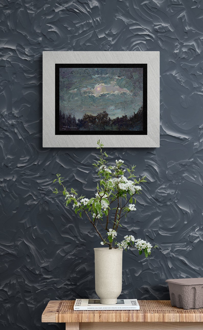 May include: An oil painting of a night landscape with a silver frame hangs on a textured gray wall. A vase with white flowers sits on a wooden table. The painting depicts a moonlit sky over a forest.