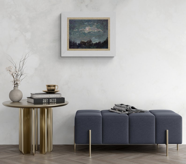 May include: A framed landscape painting depicts a night scene with a moon and trees. A small, round, gold-colored side table holds books, a teacup, and a vase with branches. A blue upholstered bench with gold legs is also present.