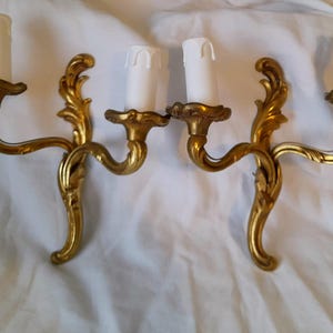 May include: A pair of ornate gold wall sconces, each with two white candle-shaped light fittings. The sconces feature a curved design with decorative flourishes. The background is white fabric.