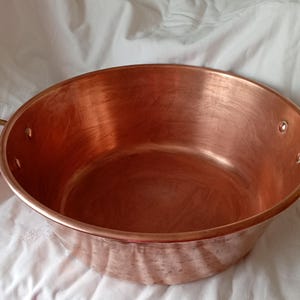 May include: A large, round, polished copper basin with a wide opening and a smooth interior surface. The vessel features two curved brass handles securely attached to opposite sides with visible metal rivets, creating a functional design.