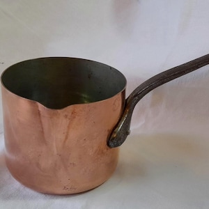 May include: A small copper saucepan with a long, dark handle. The pot has a rounded body and a shiny, reddish-brown exterior. The handle is riveted to the side of the pot.