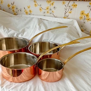 May include: A set of four copper saucepans with polished stainless steel interiors and long, slender brass-toned handles. The pans are arranged in a cluster, showcasing their metallic exterior finish and the reflective silver-toned cooking surfaces.