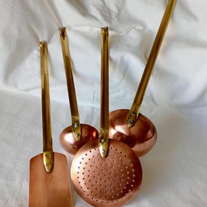 May include: A set of four kitchen utensils featuring copper heads and long brass handles. The collection includes a flat spatula, a round skimmer with perforated holes, and two deep ladles of varying sizes, all secured with visible metal rivets.