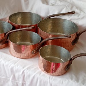 May include: A set of five hammered copper saucepans with silver-toned interiors and dark brown metal handles. Each pan features a rounded body with a textured exterior surface and a single long handle attached with rivets to the side of the vessel.