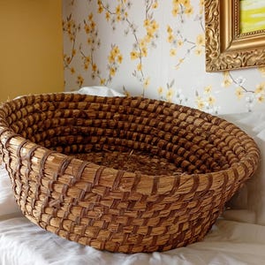 May include: An oval woven basket in natural brown tones. The basket is made from tightly woven straw, with a robust build. The rim is reinforced with a darker brown material. The empty basket is displayed on a white surface.
