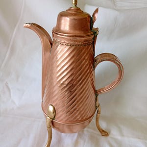 May include: A copper coffee pot featuring a diagonal ribbed texture on the body, a hinged lid with a small finial, a curved spout, and a C-shaped handle. The vessel is supported by three slender brass legs with a metallic finish and patina.