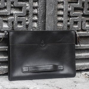 May include: Black leather clutch with a handle and zipper closure. The clutch features a small anchor emblem on the front. The bag is rectangular and sits on a concrete surface in front of a metal gate.