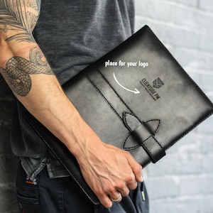 May include: Black leather portfolio with a decorative clasp. The portfolio has the text "CLIENTSIDE PM PROJECTS PROCESS PROGRAM" and a place for a logo. The portfolio is held against a grey brick wall.