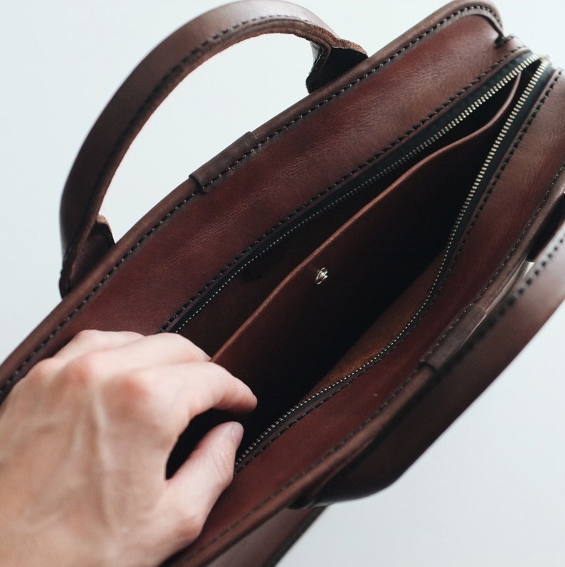 May include: A brown leather briefcase with a zipped pocket and a snap closure. The briefcase is open and the inside is visible.