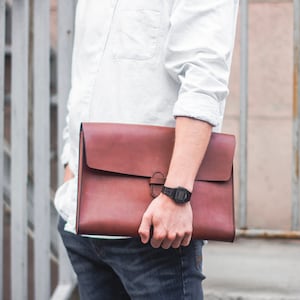 May include: A brown leather portfolio with a flap closure and a strap. The portfolio is held by a person wearing a white shirt and blue jeans.