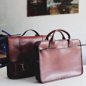 May include: Two brown leather briefcases. The larger briefcase has a brass clasp and the smaller briefcase has the initials "SRC" embossed on the front.