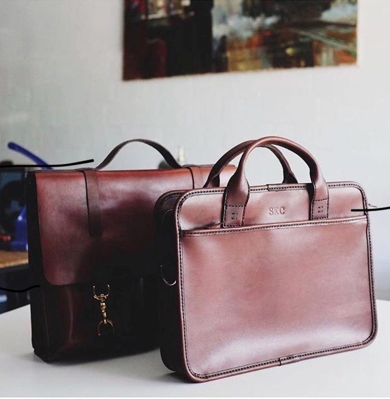 May include: Two brown leather briefcases. The larger briefcase has a brass clasp and the smaller briefcase has the initials "SRC" embossed on the front.