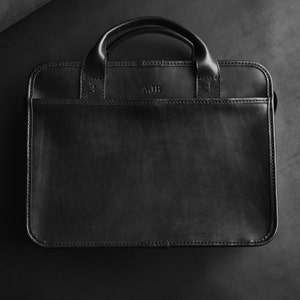 May include: A black leather briefcase with a top handle and a flap closure. The briefcase has a monogrammed initial "AJB" on the front.