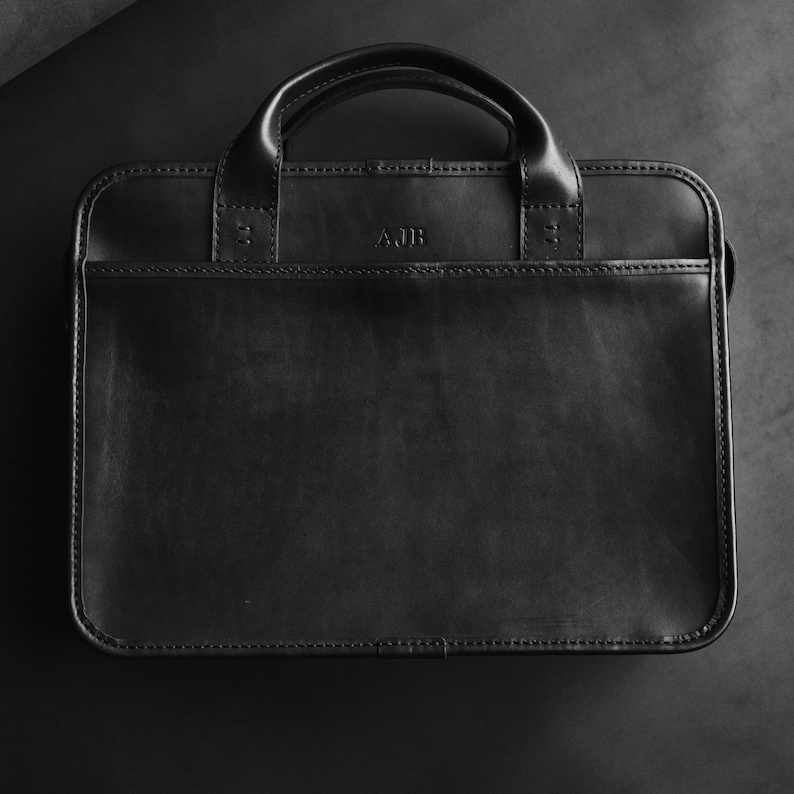 May include: A black leather briefcase with a top handle and a flap closure. The briefcase has a monogrammed initial "AJB" on the front.