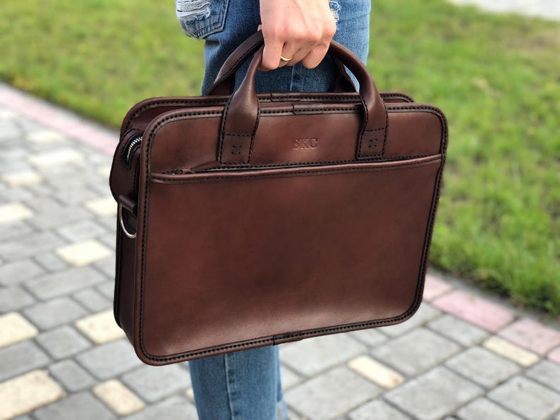 May include: A brown leather briefcase with a top handle and a shoulder strap. The briefcase has a zipped closure and a small pocket on the front. The brand name "SKC" is embossed on the front of the bag.