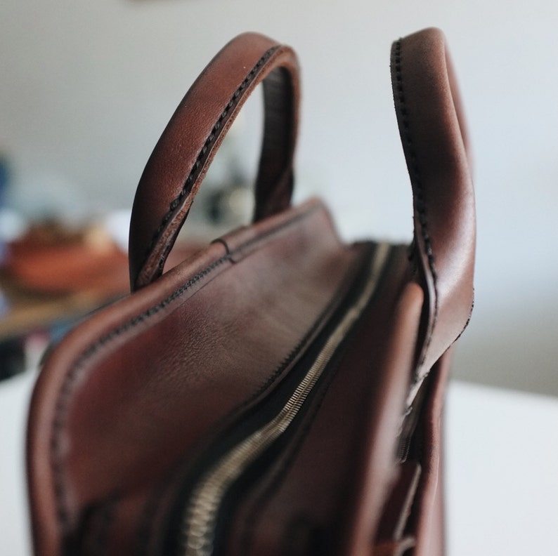 May include: A brown leather briefcase with a zipper closure and two handles. The briefcase has a stitched design and a textured surface.