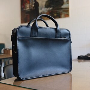 May include: A black leather briefcase with a top handle and a zipped closure. The briefcase is sitting on a wooden table.