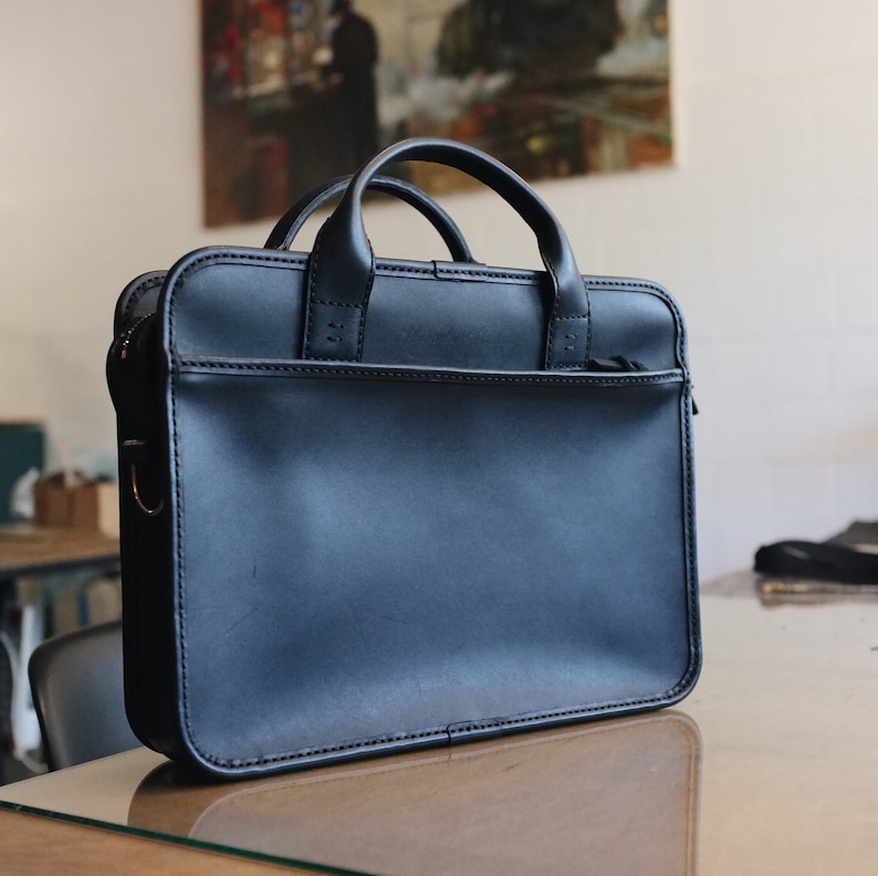 May include: A black leather briefcase with a top handle and a zipped closure. The briefcase is sitting on a wooden table.