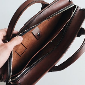 May include: A brown leather briefcase with a zipped closure and a small pocket inside. The briefcase has two handles and is open to show the interior.
