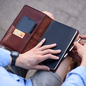 May include: A brown leather notebook cover with a black notebook, a pen, and a smartphone in a pocket. A credit card is visible in the pocket.