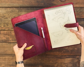 Könnte beinhalten: Ein Portfolio aus burgunderfarbenem Leder mit einem integrierten Fach für ein Tablet und einem Stifthalter. Das Portfolio ist geöffnet und zeigt ein Notizbuch mit handgezeichneten Skizzen.