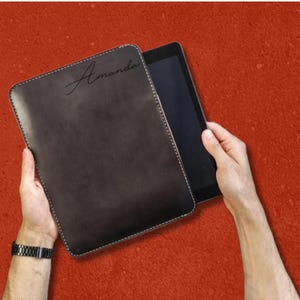 Pode incluir: Uma capa para tablet castanho-escura com cantos arredondados e costuras brancas ao longo das bordas. O nome Amanda está gravado em letra cursiva no canto superior esquerdo. A capa está entreaberta, revelando um ecrã de tablet preto.