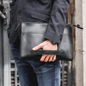 May include: A black leather clutch with a handle and a zipper closure. The clutch is rectangular and has a pocket on the front. The person is wearing a navy blue blazer and blue jeans.