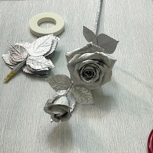 May include: A silver paper rose with a silver paper bud and leaves. The rose is on a white textured surface with a roll of tape and a pair of red handled scissors.