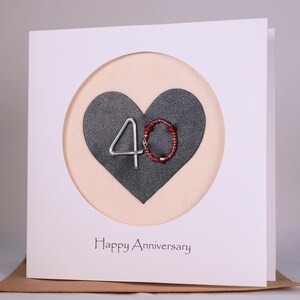 May include: A white card with a round cutout featuring a black heart with a silver wire number "40" and red beads. The text "Happy Anniversary" is printed below the heart.