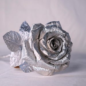 May include: A single silver rose with a textured finish. The rose is lying on a white surface.
